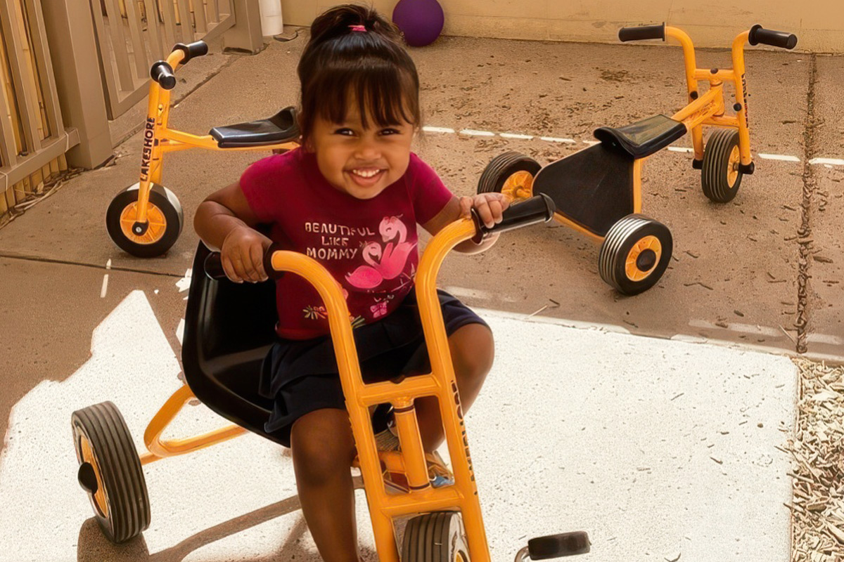 Smiling toddler riding a yellow tricycle outdoors at ElevateChild childcare in Scottsdale, AZ.